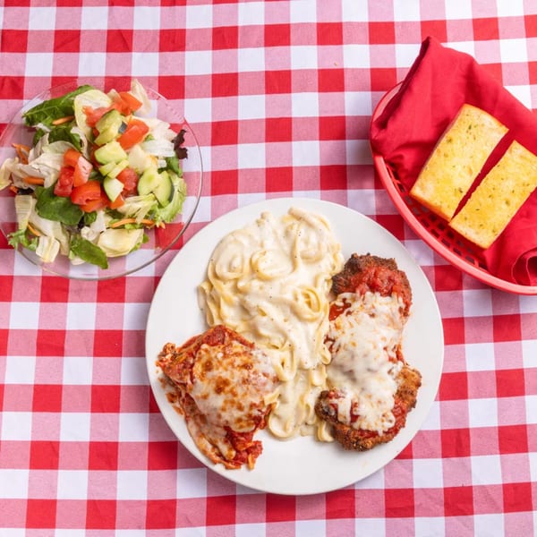 Italian dinner with lasagna, chicken parmesan, fettuccine alfredo, salad, and garlic bread on a red checkered tablecloth.