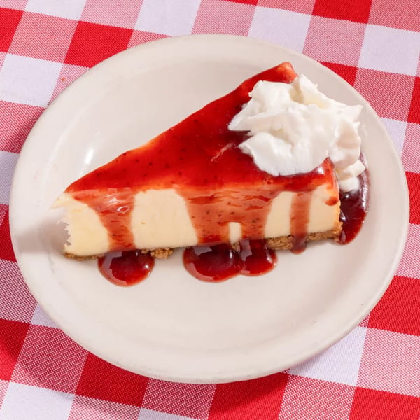 A slice of cheesecake with strawberry topping and whipped cream on a white plate over a red and white checkered tablecloth.
