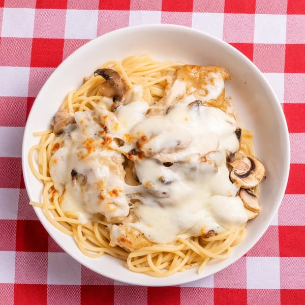 Spaghetti topped with chicken, melted mozzarella cheese, and mushrooms in a white bowl on a red and white checkered tablecloth.
