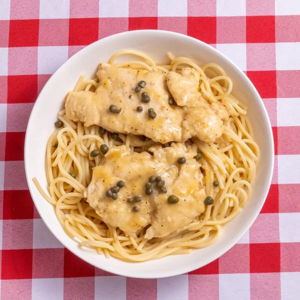Chicken piccata with capers and spaghetti in a white bowl on a red and white checkered tablecloth.