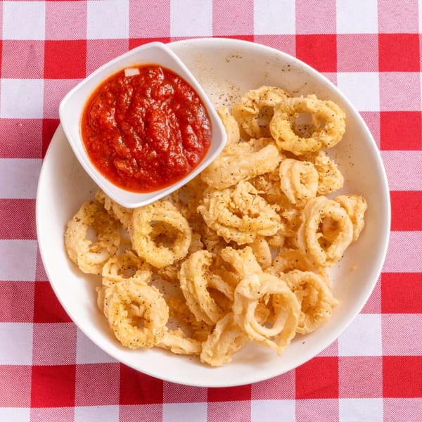 Crispy fried calamari rings with a side of red marinara sauce on a white plate over a red and white checkered tablecloth.